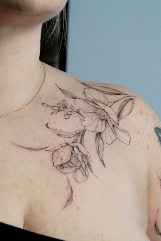 Rozbudowany i niezwykle delikatny tatuaż kwiatowy na ramieniu i obojczyku, wykonany w technice cienkiej linii (fine line) z subtelnym cieniowaniem. Projekt autorstwa Olgi (@olga_sankova_tattoo) z krakowskiego studia Fineline Tattoo Gallery. Kompozycja charakteryzuje się lekkością i płynnością, idealnie dopasowując się do kobiecej anatomii.