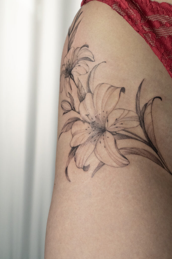 An impressive floral tattoo featuring lilies, placed on the hip and thigh, executed in the fine line technique with subtle shading. Designed and tattooed by Olga (@olga_sankova_tattoo) at Fineline Tattoo Gallery in Krakow. The composition is characterized by its delicacy and smooth tonal transitions, perfectly following the body's natural curves.