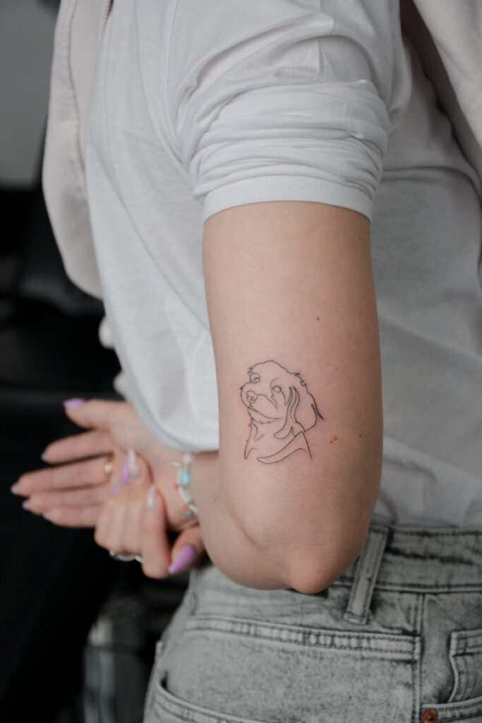 Minimalistyczny tatuaż przedstawiający portret psa, wykonany techniką jednej ciągłej linii (single line) przez Olgę (@olga_sankova_tattoo) w krakowskim studiu Fineline Tattoo Gallery. Subtelna kompozycja na ramieniu, która w nowoczesny sposób upamiętnia pupila, łącząc prostotę z precyzją stylu fine line.