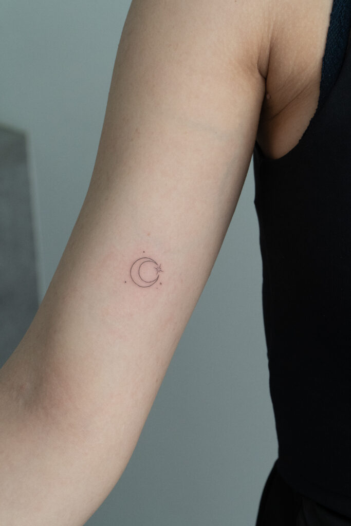 A minimalist crescent moon and star tattoo, executed with an extremely fine line. Created by Olga (@olga_sankova_tattoo) at Fineline Tattoo Gallery in Krakow. A perfect example of a "tiny tattoo" on the arm, combining precision with delicate celestial symbolism.
