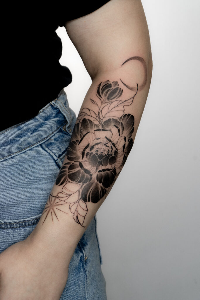 Artystyczny tatuaż na przedramieniu przedstawiający pionię w połączeniu z motywem księżyca i gwiazdy. Praca wykonana przez Olgę (@olga_sankova_tattoo) w krakowskim studiu Fineline Tattoo Gallery. Projekt charakteryzuje się mocnym kontrastem między głęboką czernią wypełnień a niezwykle delikatnymi liniami, tworząc unikalną kompozycję botaniczną.