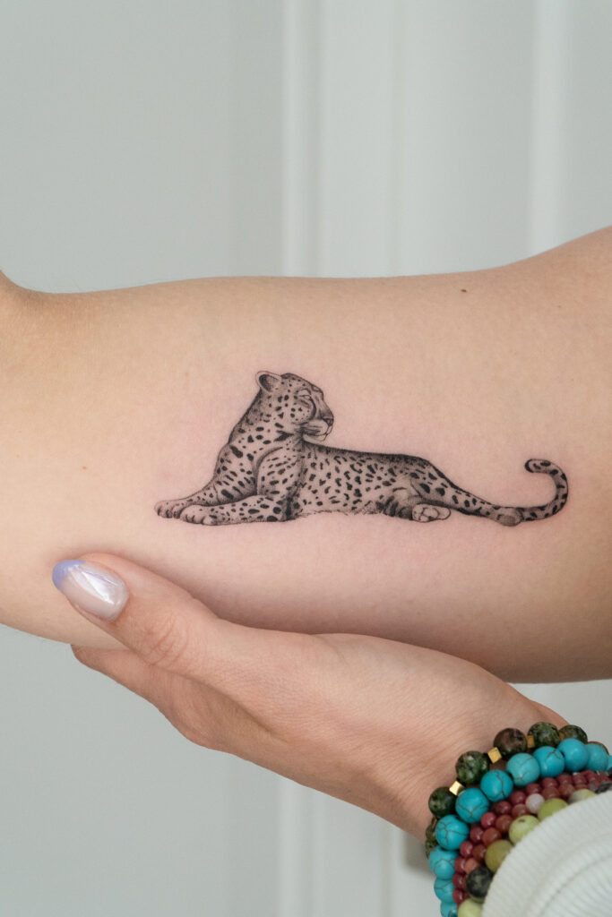 Szczegółowy tatuaż czarno-szarego lamparta odpoczywającego na przedramieniu, wykonany przez tatuatorkę Olgę Sankovą w Fineline Tattoo Gallery. Projekt wyróżnia się precyzyjnym cieniowaniem i delikatnymi liniami, które podkreślają charakterystyczne cętki zwierzęcia oraz jego spokojną, zrelaksowaną pozę. Zdjęcie ukazuje umiejscowienie tatuażu na skórze oraz dłoń delikatnie podtrzymującą ramię, aby podkreślić kunszt i dokładność wykonania.